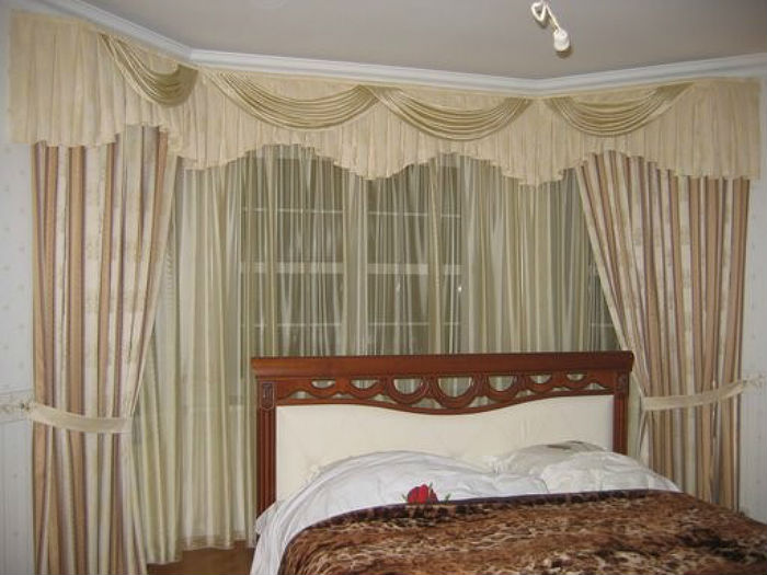 Небольшое пространство спальни лучше оформить угловыми шторами в пастельных тонах, так интерьер не будет казаться перегруженным и тяжелым